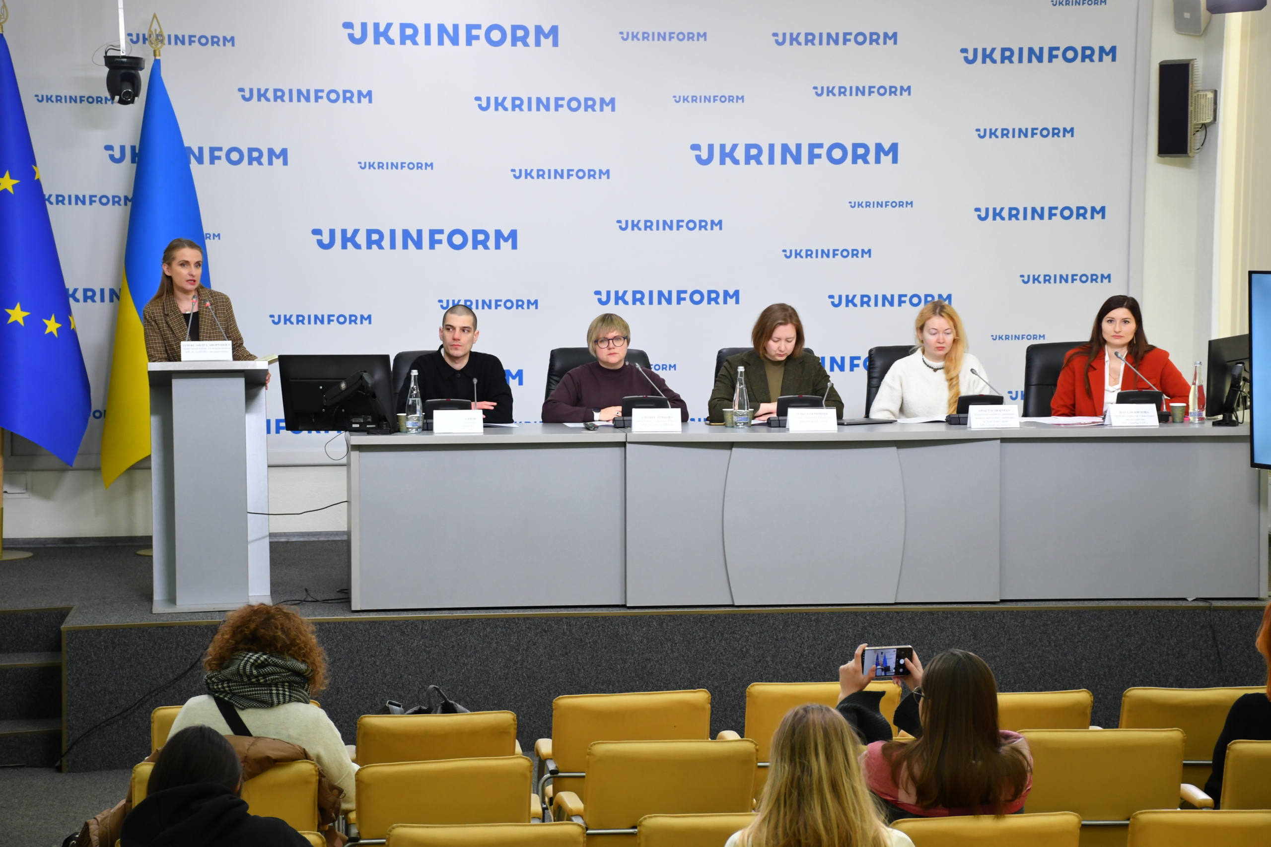 A panel of six people sits at a long gray desk during a press conference in front of a backdrop featuring the Ukrinform logo. One woman stands at a lectern to the left of the European Union and Ukrainian flags, while audience members in yellow chairs listen and record the event on smartphones.