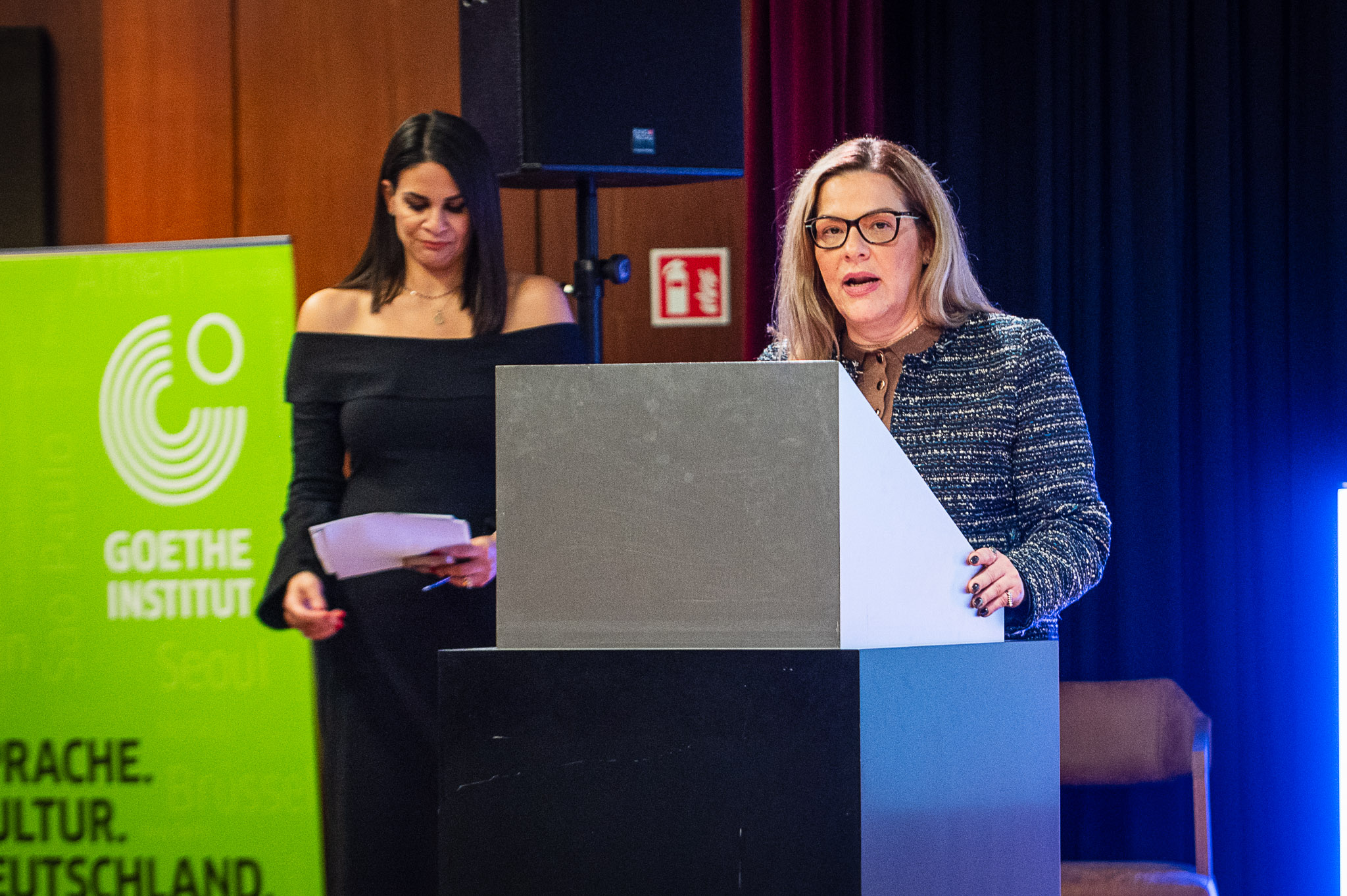 A woman with long blonde hair and glasses speaks from behind a light-grey podium at a Goethe-Institut event. In the background, another woman, Tina Michailidou, journalist, SKAI TV; Founder, GR8Communication, in a black off-the-shoulder dress stands holding papers, while a green banner with the "Goethe-Institut" logo is visible on the left.