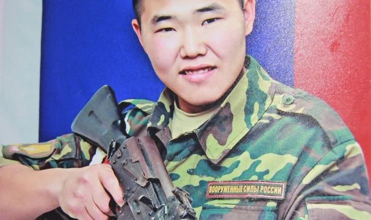 A commemorative-style portrait shows a Russian soldier with Asian features, identified as ANDREI ANDREEV, wearing a camouflage uniform and a black marine beret. He is smiling and holding a Kalashnikov assault rifle against a background of the Russian flag.