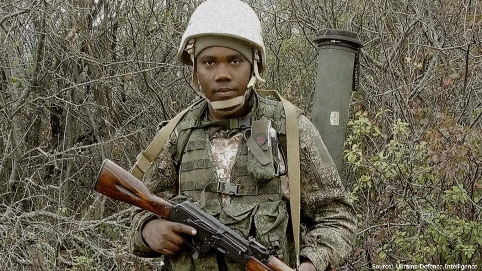 A man of African descent in a green camouflage military uniform and a white helmet stands in front of dense, leafless thickets while holding an assault rifle. He has a military backpack with a green cylindrical tube attached to the side, and a "Ukraine Defence Intelligence" source credit appears in the bottom-right corner.