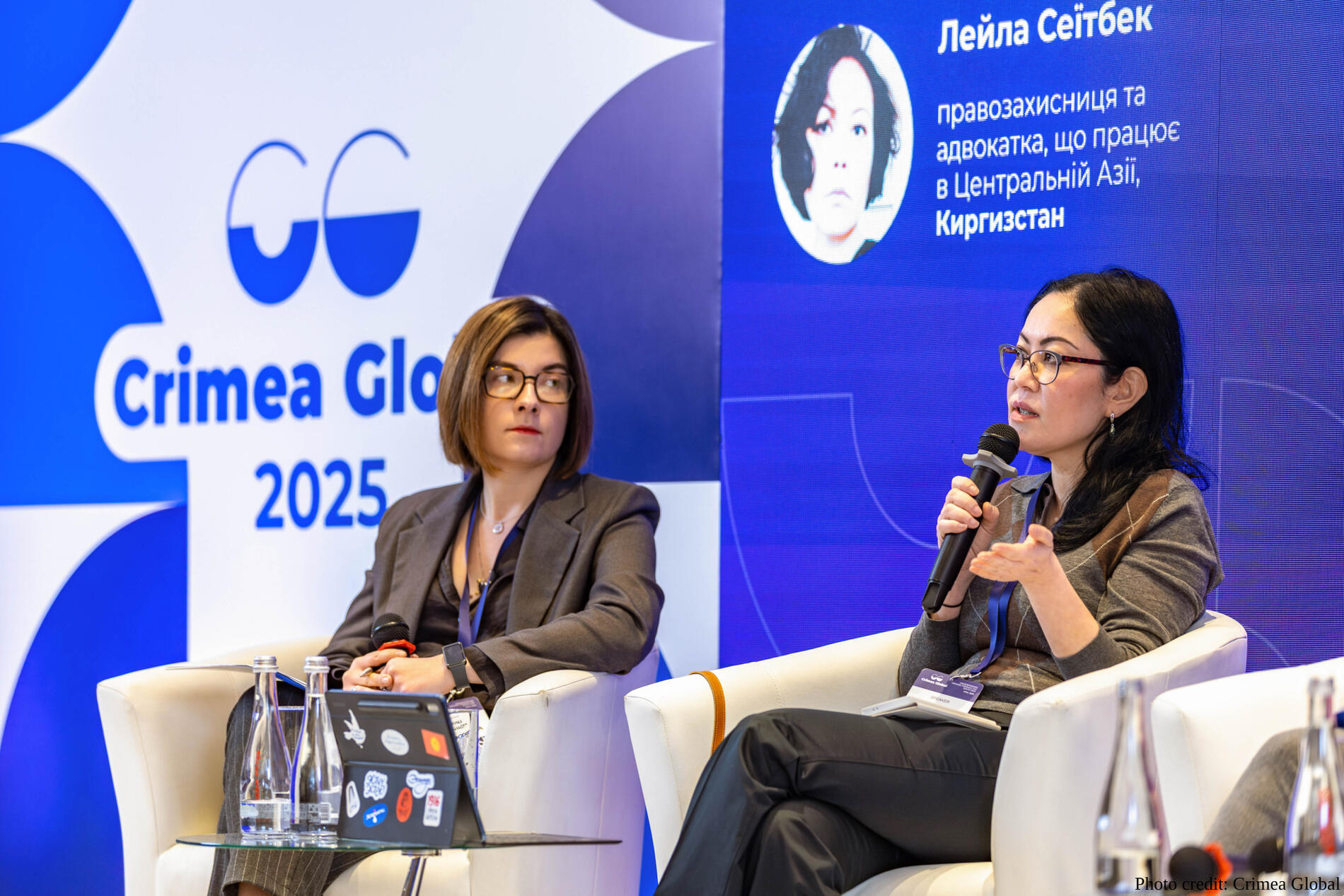 Human rights activist Leila Seiitbek is seated in a white armchair during a panel discussion. She wears glasses and a gray argyle sweater, holding a microphone and gesturing while speaking. Behind her is a blue digital backdrop with "Crimea Global 2025" and another participant.