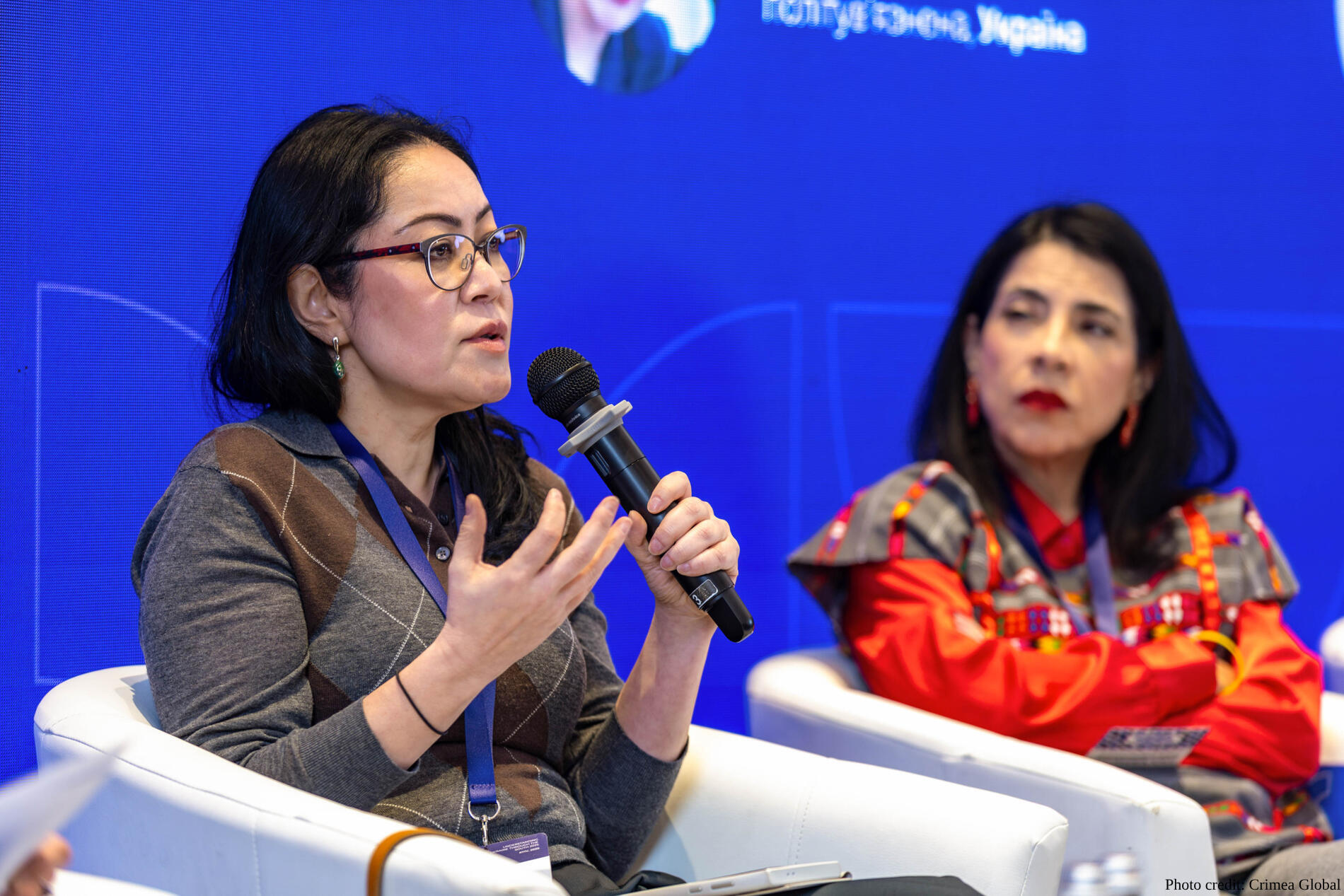 Human rights activist Leila Seiitbek from Kyrgyzstan is pictured sitting in a white armchair during a panel discussion. She wears glasses and a gray argyle sweater, holding a microphone in her right hand and gesturing with her left while speaking. Another woman in a red and black traditional patterned garment is seated behind her against a blue digital backdrop.