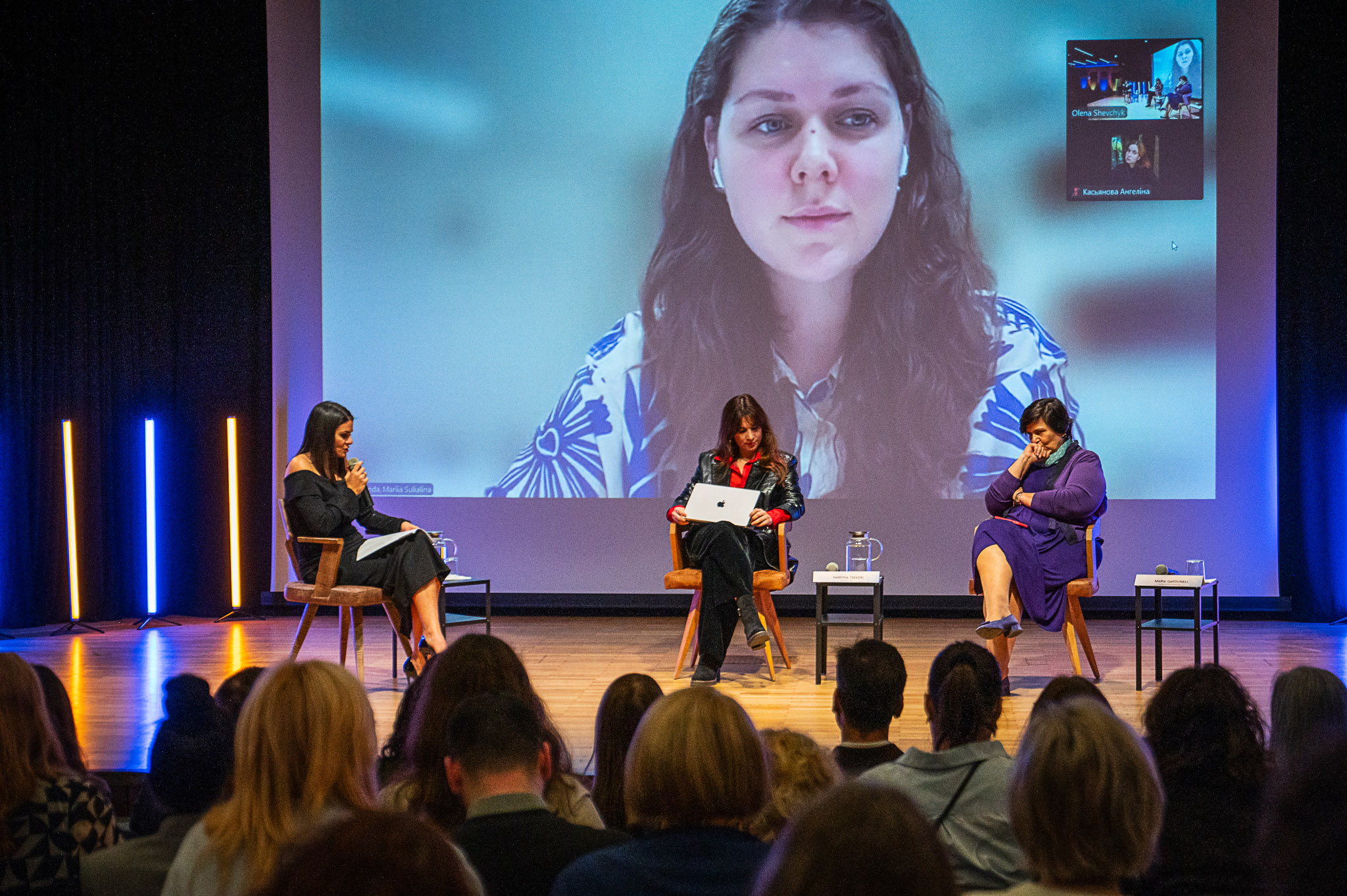 Human rights activist Maria Sulyalina is shown on a large video screen, participating remotely in a panel discussion. In the foreground, three women are seated on a stage: one speaks into a microphone, one works on a laptop, and one listens attentively before an audience.