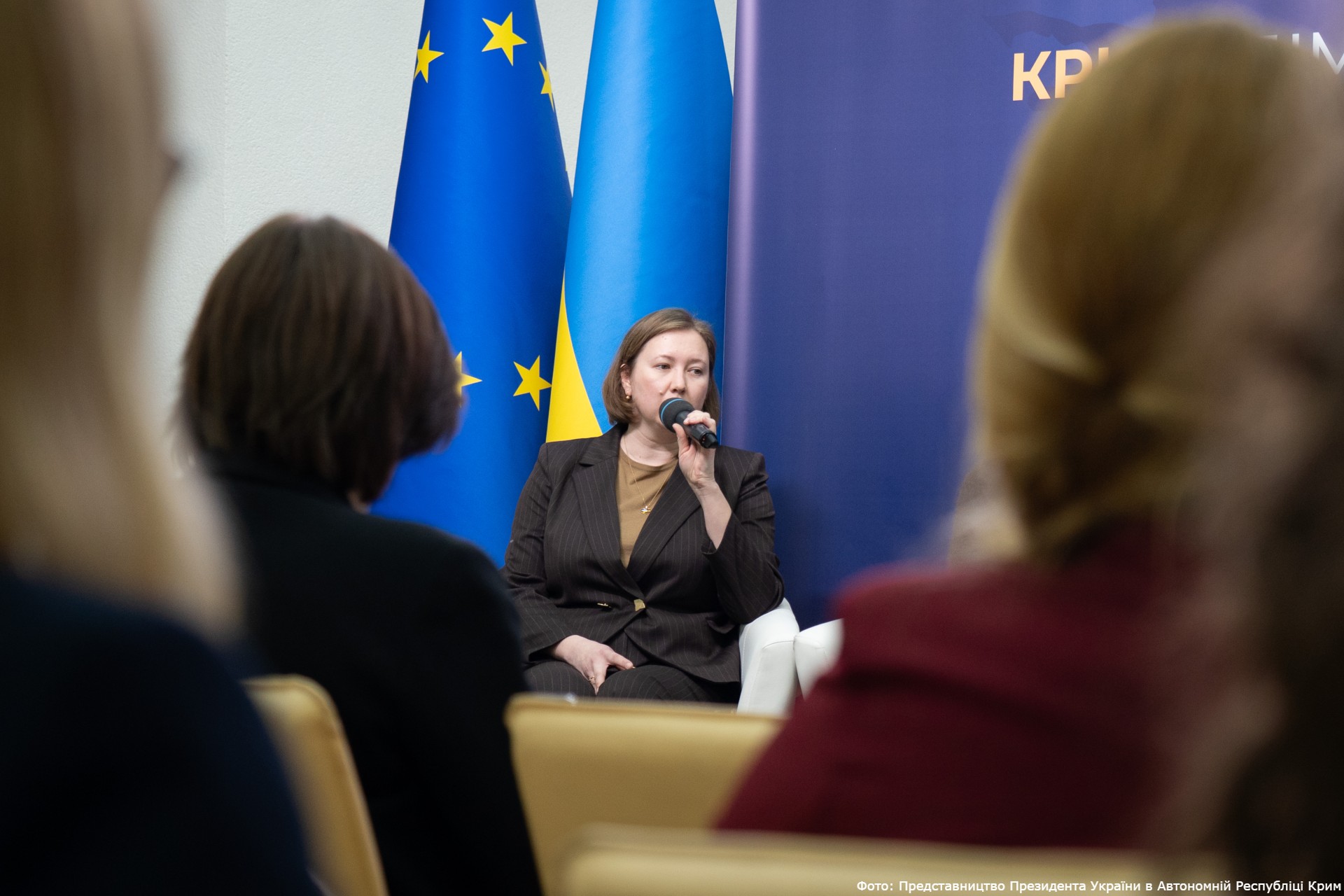 Human rights defender Olha Skrypnyk, head of the Crimean Human Rights Group, speaks into a microphone at a forum in Kyiv. She is seated between the flags of the European Union and Ukraine. The event, titled "CRIMEA: The Home We Are Fighting For," was held on February 26, 2026, to mark the 12th anniversary of resistance to the Russian occupation of the peninsula.