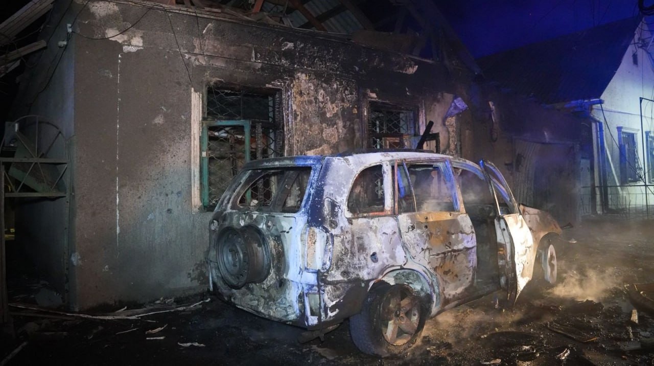 The charred, smoldering frame of a white SUV with its doors open sits in front of a heavily damaged, soot-covered building at night. The logo for the State Emergency Service (SES) of Ukraine in the Odesa region is positioned in the upper-left corner of the image.