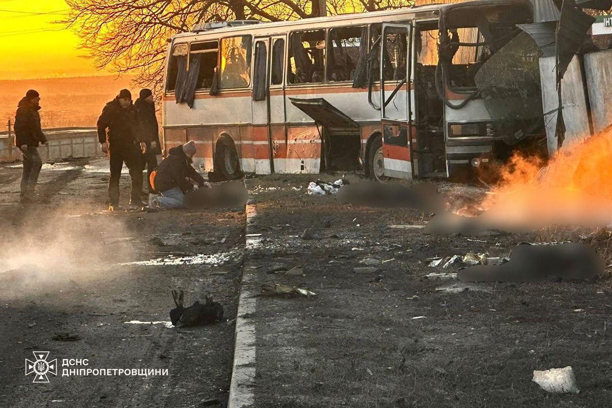 Emergency personnel examine a burned and shrapnel-damaged company bus on a roadside near Pavlohrad following a drone strike. Several blurred shapes on the ground indicate the presence of victims, and a fire burns in the background near a structure, with the DSNS Dnipropetrovsk logo in the bottom left.