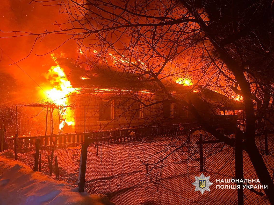 A single-story residential building is engulfed in a bright orange fire at night, with flames rising through the roof. Skeletal tree branches are silhouetted against the intense glow, and the logo for the National Police of Ukraine is positioned in the bottom-right corner.