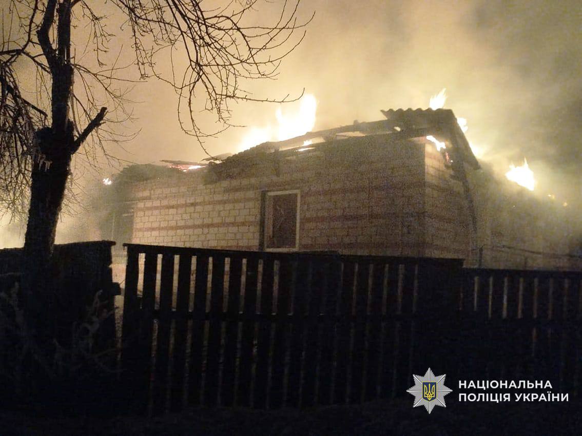 A brick building smolders after a fire, with individual pockets of flame still visible through the destroyed roof against the night sky. A dark wooden fence stands in the foreground, and the logo for the National Police of Ukraine is located in the bottom-right corner.