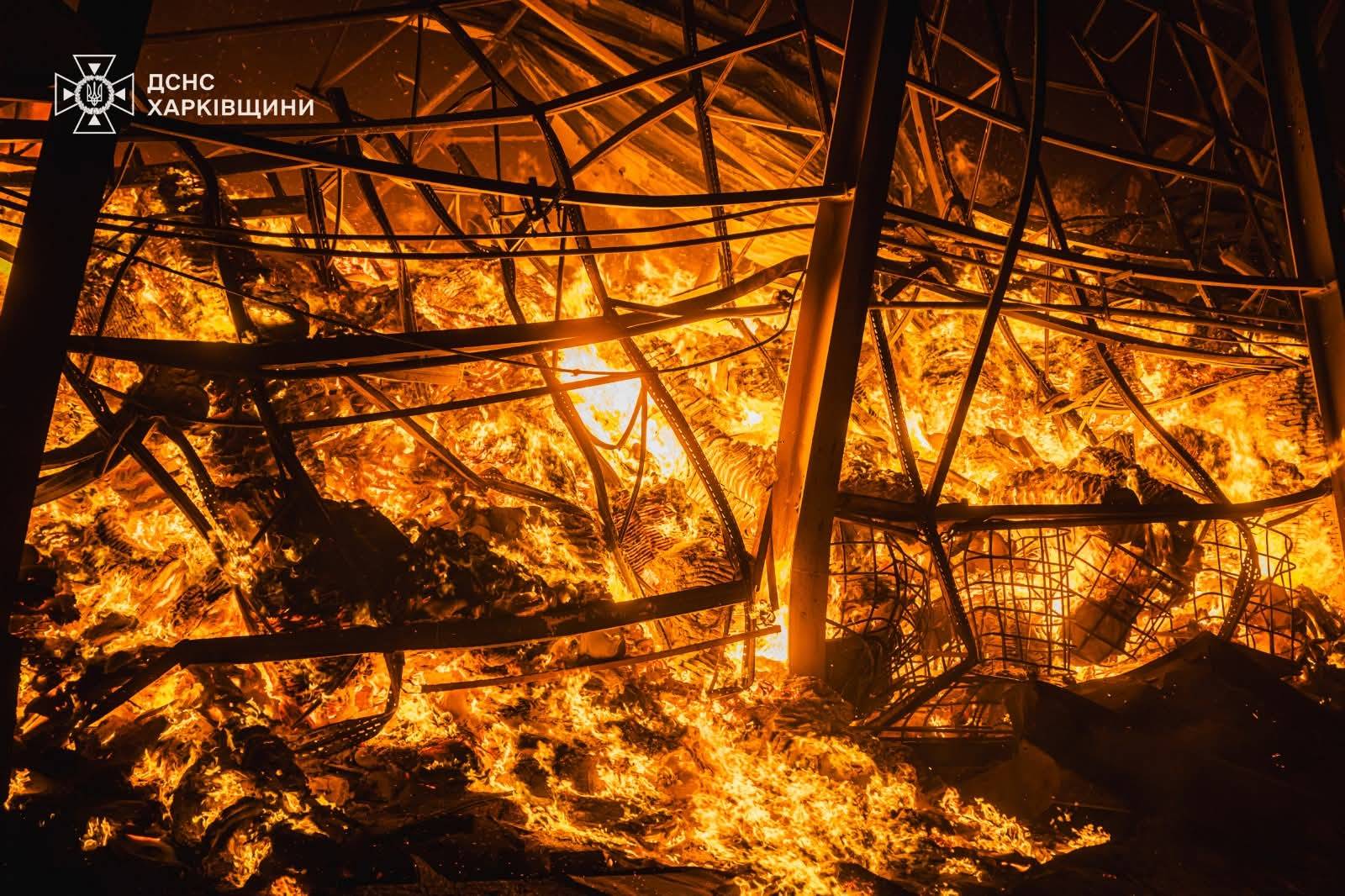 Large, orange flames consume a tangled web of metal structural beams and debris during a night fire. The logo for the State Emergency Service (SES) of Ukraine in the Kharkiv region is visible in the upper-left corner of the frame.
