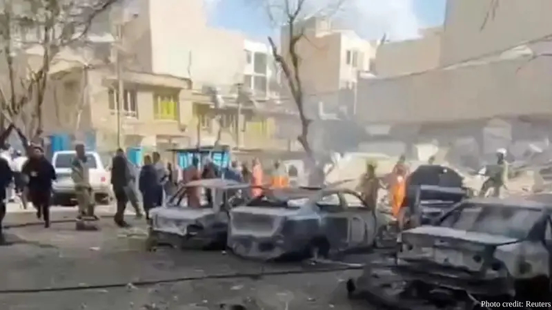 The blurred aftermath of a major military strike in an urban area of Tehran shows thick smoke rising behind multi-story residential and commercial buildings. In the foreground, several civilian cars are completely charred and destroyed, and debris is scattered across the street as people run through the scene. This image documents the joint U.S.-Israeli preemptive operation "Lion's Roar" launched against the Iranian regime on February 28, 2026.