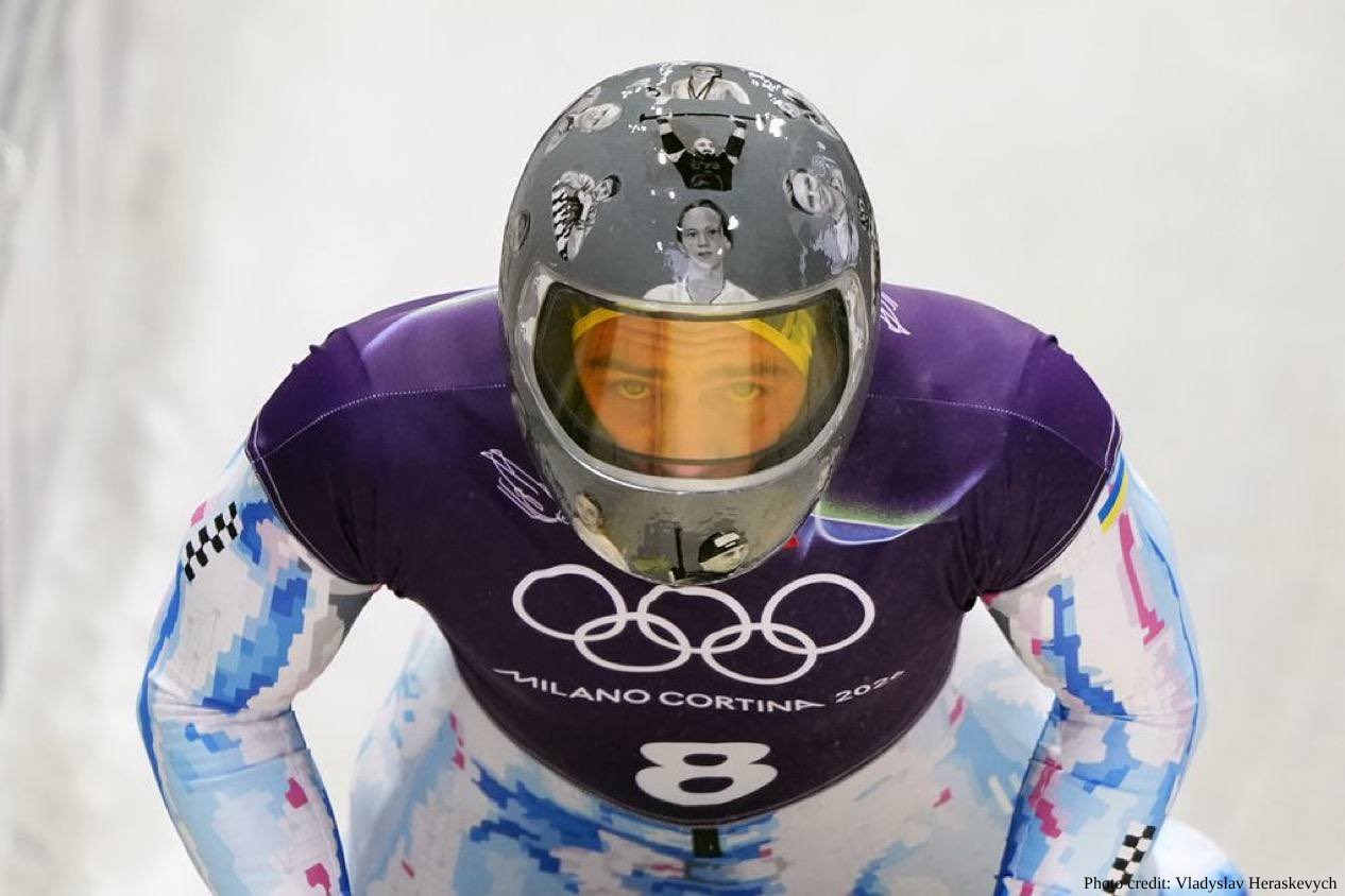 A skeleton athlete Vladyslav Heraskevych wearing a purple and white racing suit with the Milano Cortina 2026 Olympic logo looks forward from a crouched position. The athlete’s grey helmet is decorated with black-and-white portraits of several individuals.