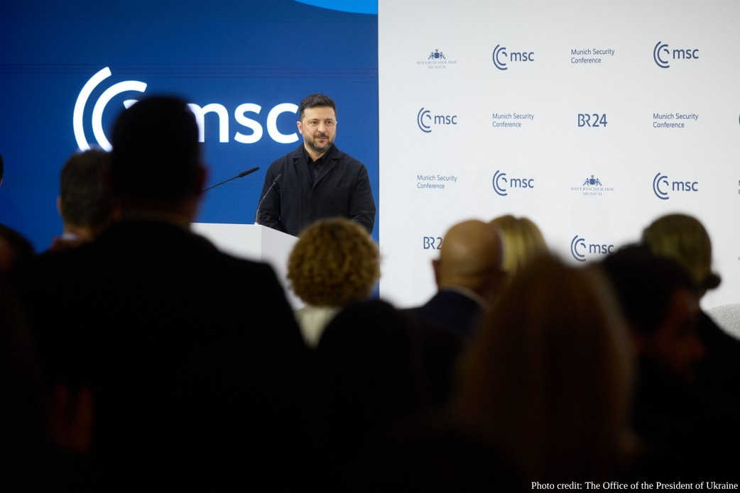 Ukrainian President Volodymyr Zelenskyy speaks from behind a white podium at the Munich Security Conference, with the "msc" logo prominent on the blue and white backdrop. He is viewed from behind a crowd of attendees in a dimly lit room.