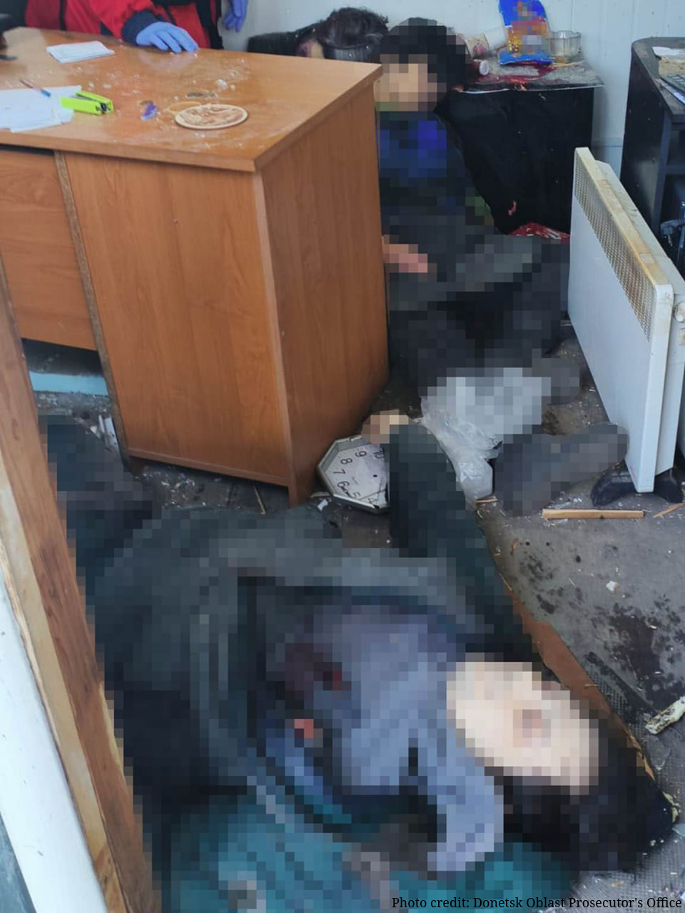 The interior of a damaged room shows the blurred remains of victims lying on a floor covered in dust and debris. A light-colored wooden desk stands on the left, and a white wall clock lies face-up on the ground with its hands stopped at approximately 8:15. A white space heater is attached to the wall in the background, surrounded by scattered papers and office supplies.