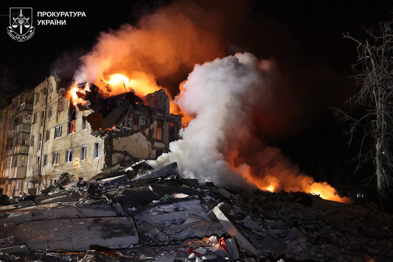 A multistory apartment building is engulfed in flames and thick white smoke at night, with a massive pile of rubble in the foreground where a section of the structure has collapsed. The Prosecutor General of Ukraine logo is visible in the upper left corner.