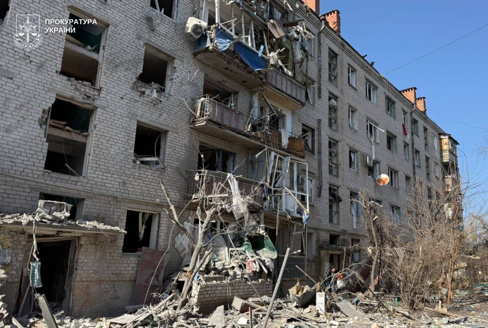 A five-story residential building shows extensive damage with shattered windows and destroyed balconies, while the ground is covered in rubble and twisted metal. The Prosecutor General's Office of Ukraine logo is featured in the top left corner.