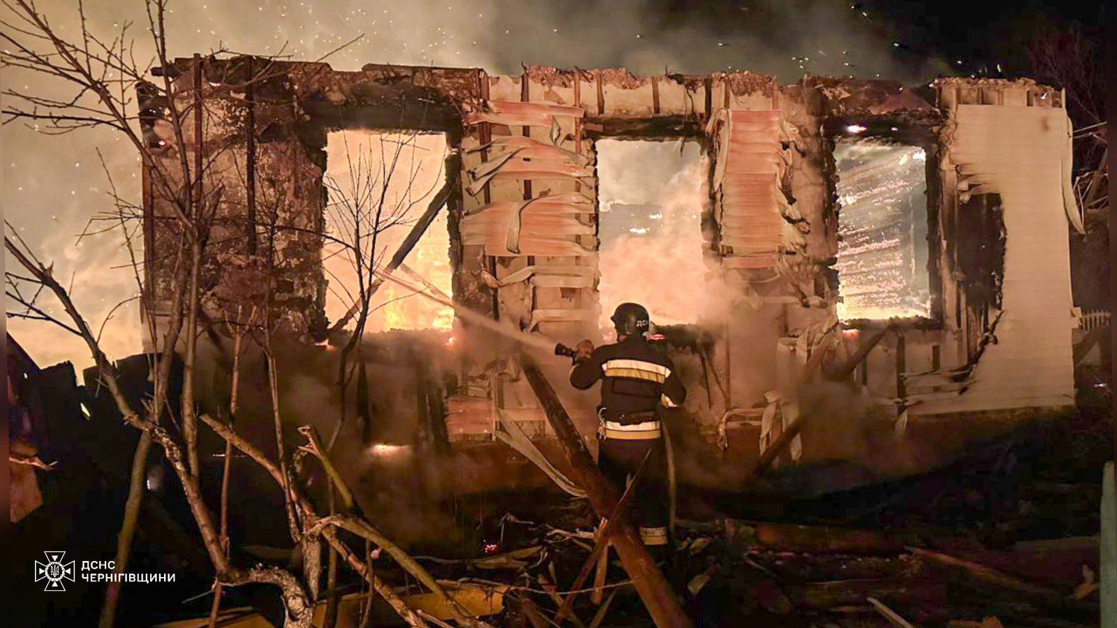 A firefighter stands with his back to the camera, directing a hose at the skeletal, flaming remains of a building at night. The State Emergency Service of Ukraine (DSNS) Chernihiv region logo is visible in the bottom left corner.
