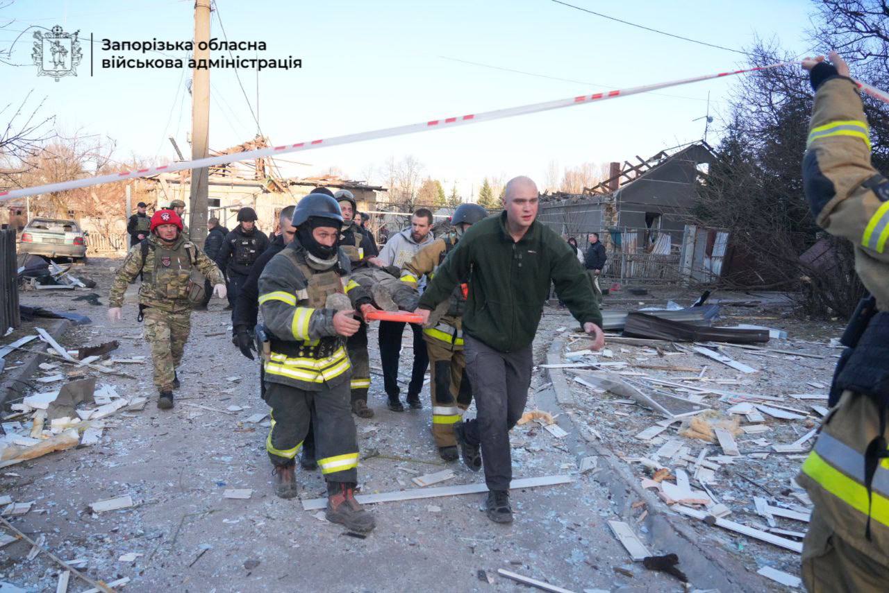 Firefighters and personnel in tactical gear carry a person on a stretcher through a yard covered in glass and debris from nearby damaged houses. The Zaporizhzhia Regional Military Administration logo is visible in the top left corner.