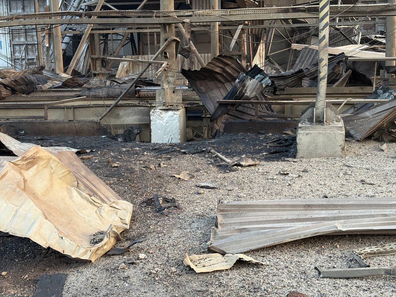 A wide shot of extensive damage at an industrial facility, showing mangled metal beams, twisted corrugated sheets, and scorched ground covered in debris. The structural framework is partially collapsed, with shattered materials strewn across the area.