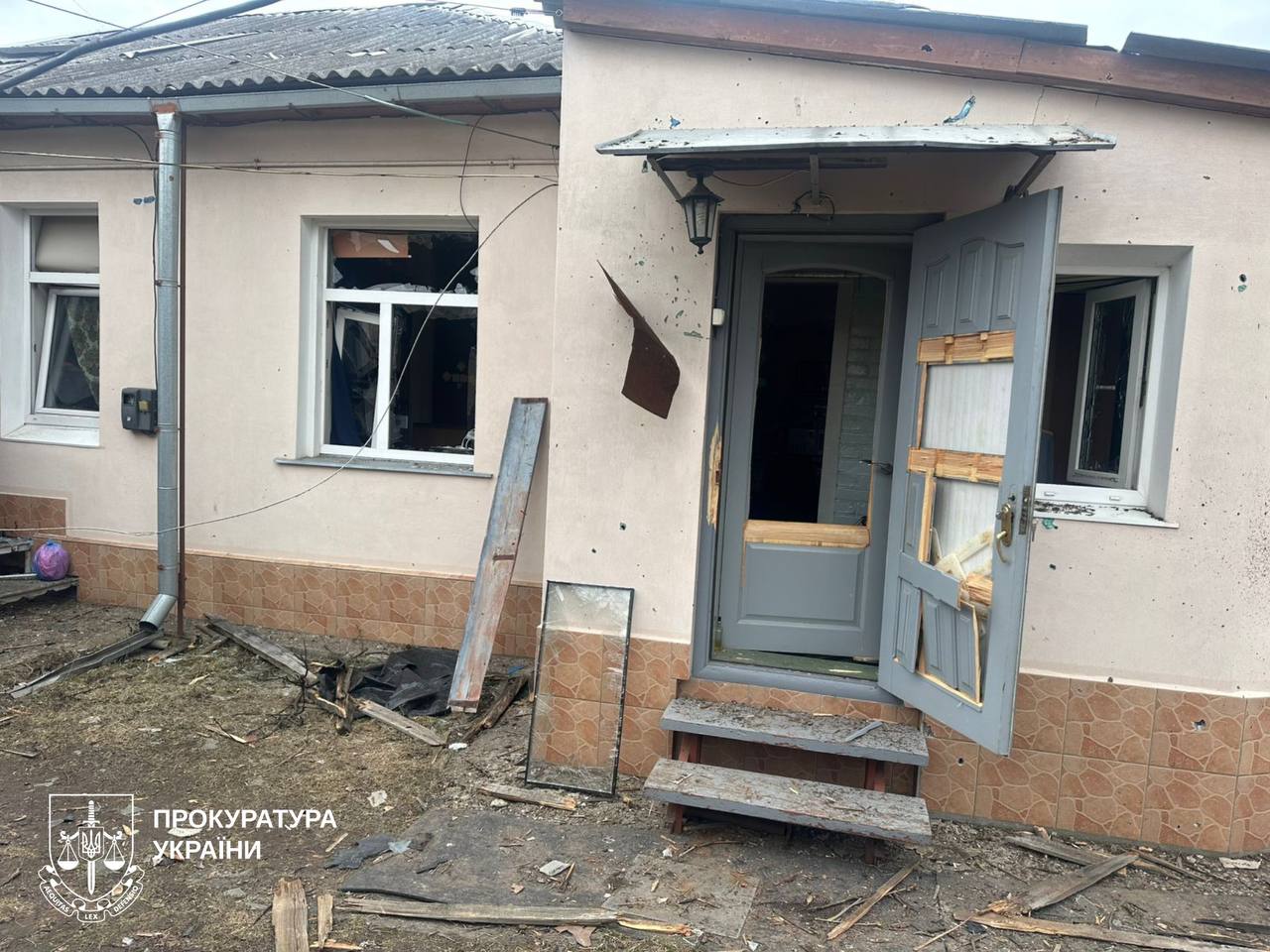 The image shows the aftermath of a Russian drone strike on a residential area, specifically a one-story private house. The exterior wall is scorched and covered in soot, with a shattered window and a damaged grey front door hanging off its frame. Debris from the roof and cladding is scattered across the yard. The bottom-left corner features the official watermark of the Prosecutor's Office of Ukraine.
