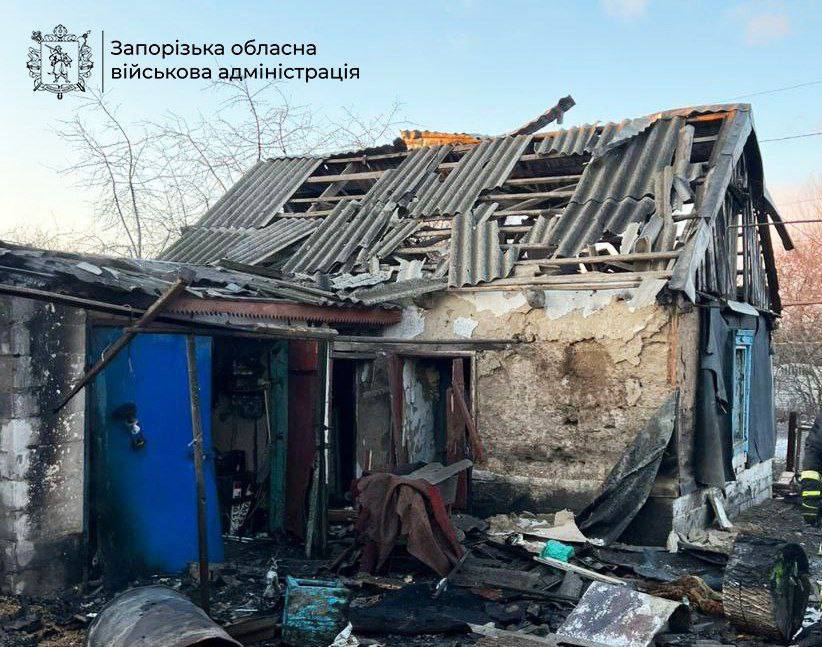 A small, one-story residential house is in ruins following an attack. The slate roof is largely destroyed, with shattered tiles and exposed wooden beams. The walls are crumbling, and the interior is filled with debris. A bright blue metal door stands partially detached amidst the wreckage of wood and personal belongings. The official watermark of the Zaporizhzhia Regional Military Administration is visible in the top-left corner.