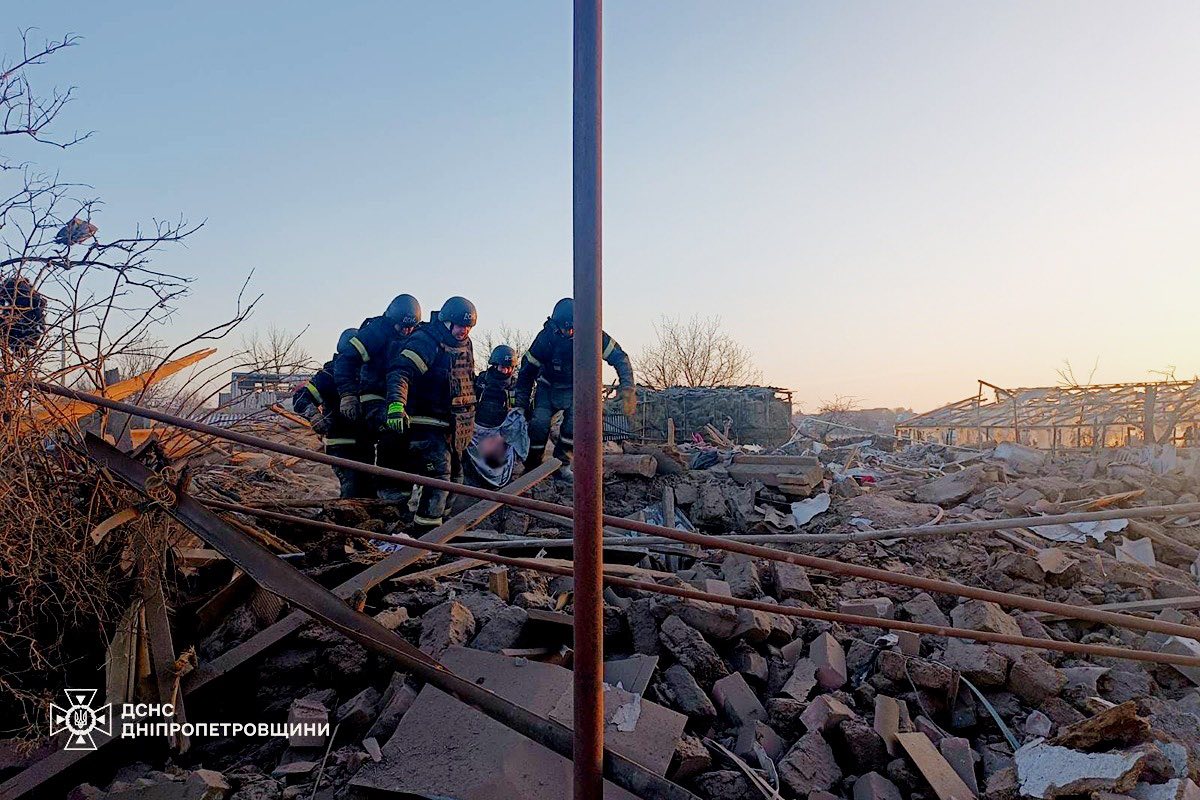 A team of State Emergency Service (SES) rescuers in dark protective gear and helmets carries a person wrapped in a grey sheet across a vast field of building debris, including bricks, concrete blocks, and twisted metal beams. The scene is captured at sunrise or sunset, with the sun low on the horizon. A thin metal pole stands in the foreground, and the logo "ДСНС ДНІПРОПЕТРОВЩИНИ" is visible in the bottom-left corner.