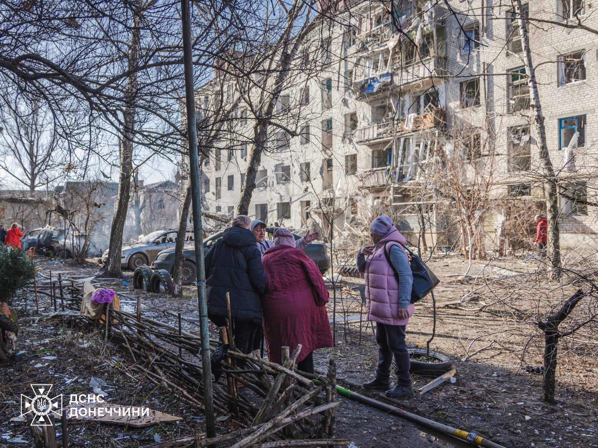 Several women stand in a debris-strewn courtyard talking, with a heavily damaged apartment building and scorched cars visible in the background. The State Emergency Service of Ukraine (DSNS) Donetsk region logo is located in the bottom left corner.