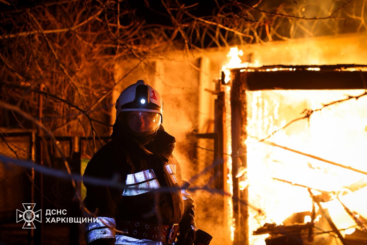 A firefighter in protective gear and a helmet with a headlamp stands in front of a structure engulfed in bright orange flames at night. The State Emergency Service of Ukraine logo for the Kharkiv region is visible in the bottom left corner.