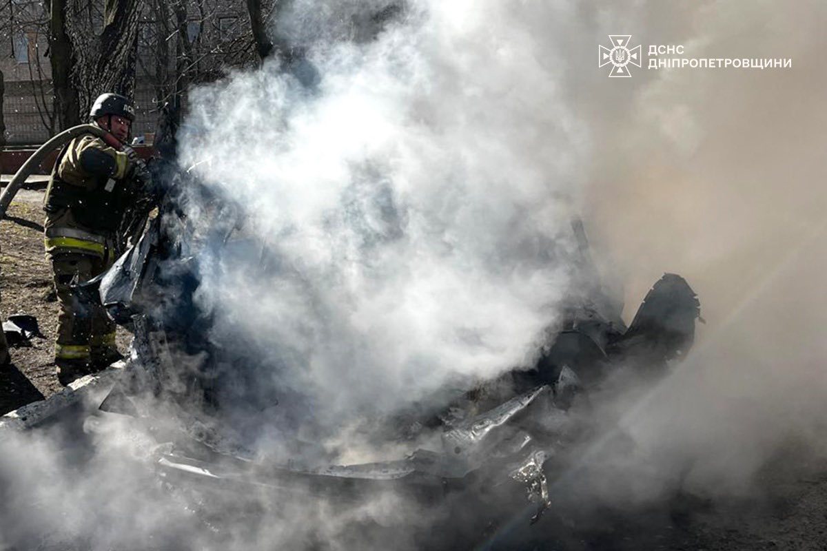 A firefighter in full protective gear aims a hose at a smoldering, unrecognizable vehicle engulfed in thick white smoke. The State Emergency Service of Ukraine (DSNS) Dnipropetrovsk region logo is visible in the top right corner.