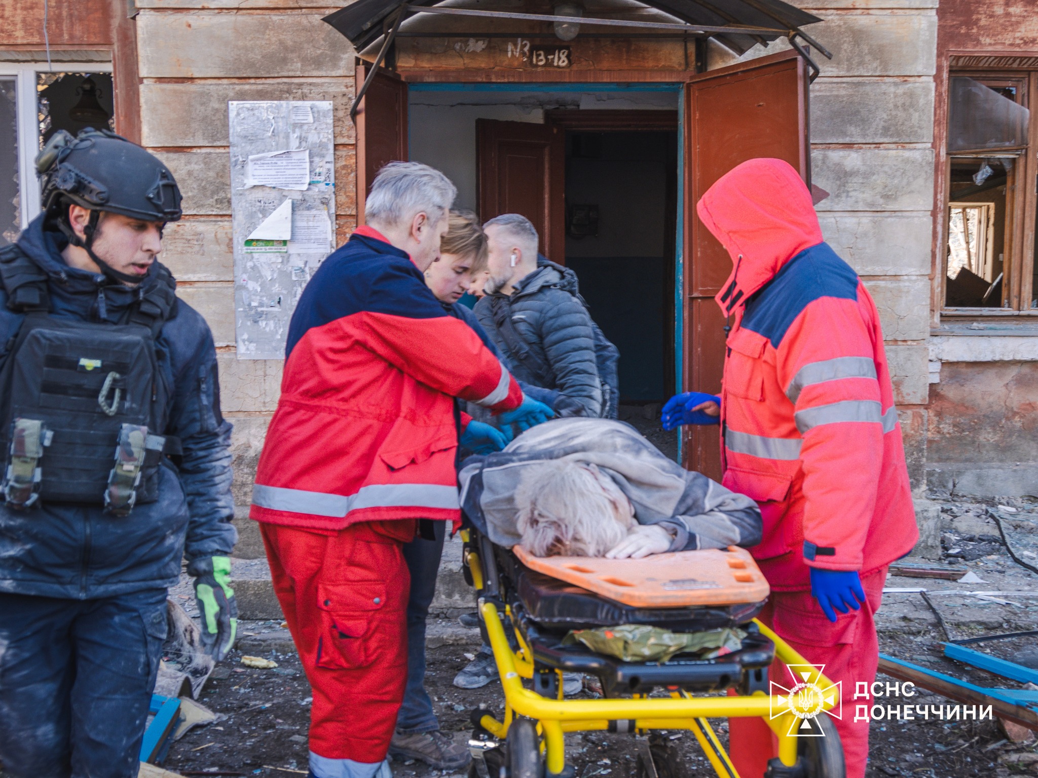 Emergency responders in red uniforms and a person in tactical gear move a victim on a yellow stretcher away from the entrance of a damaged building. The State Emergency Service of Ukraine (DSNS) Donetsk region logo is visible in the bottom right.