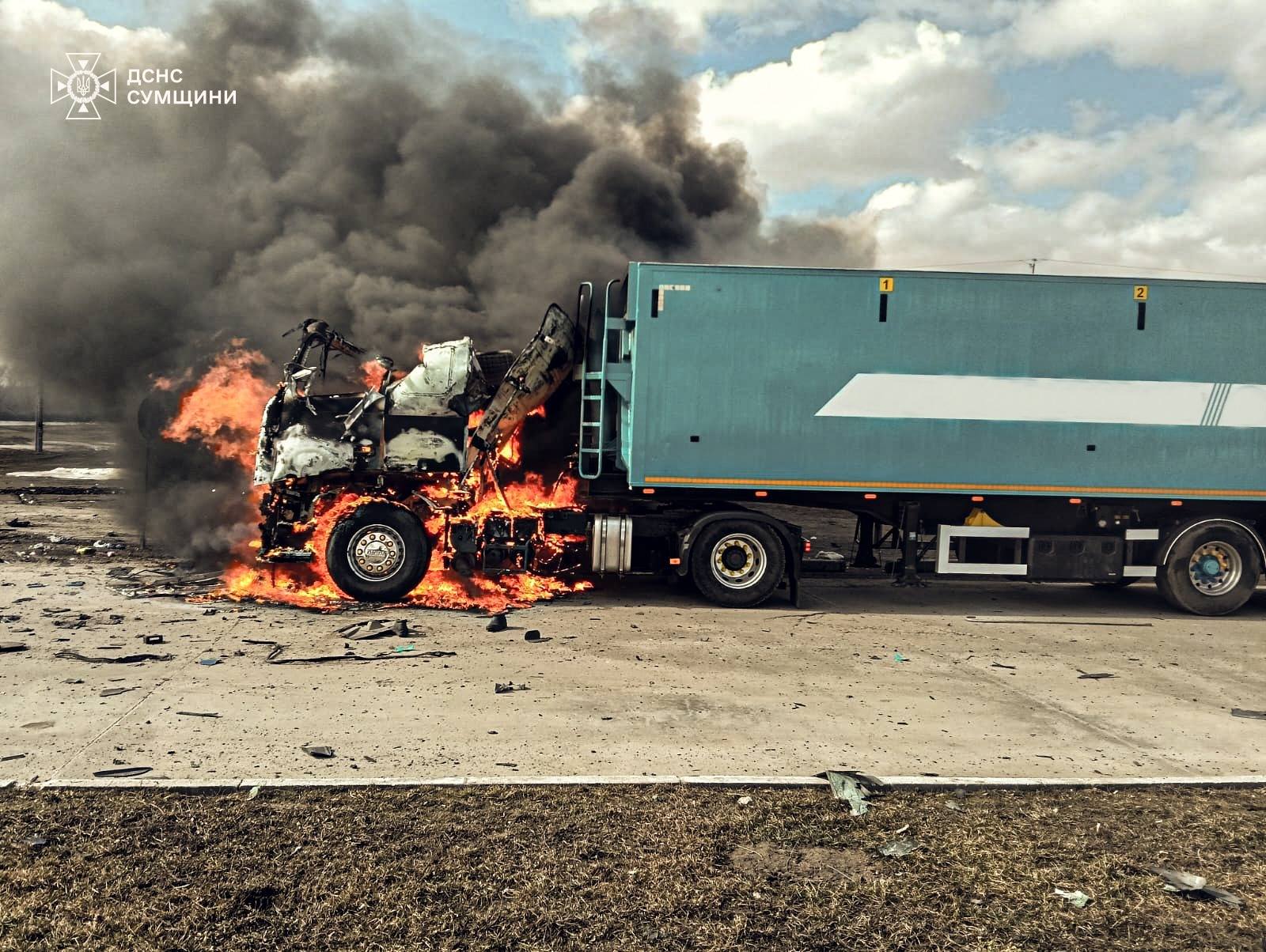 A semi-truck with a teal trailer has its cab completely engulfed in bright orange flames and thick black smoke on the side of a road. The State Emergency Service of Ukraine (DSNS) Sumy region logo is visible in the top left corner.