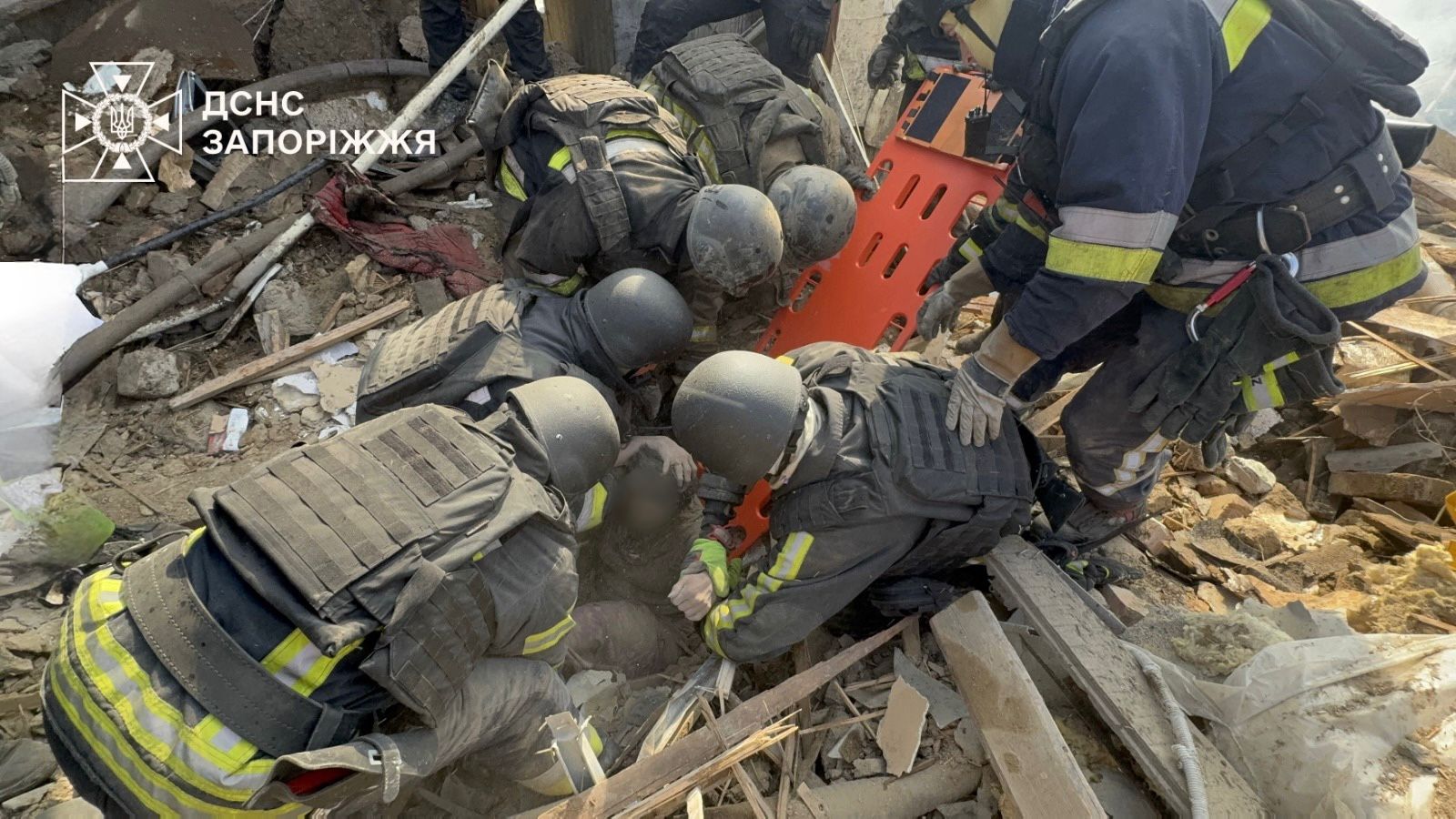 Multiple rescue workers in tactical gear and helmets carefully lift a person onto an orange stretcher amidst a large pile of destroyed concrete and wood. The State Emergency Service of Ukraine (DSNS) Zaporizhzhia logo is in the top left corner.