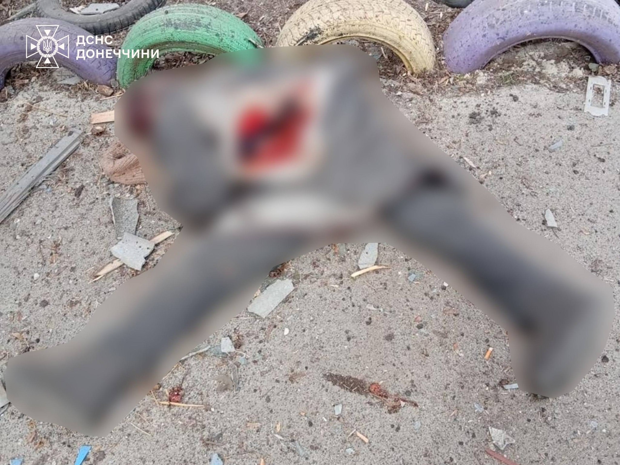 A blurred image shows a body lying on the ground near painted tires in a courtyard, with the State Emergency Service of Ukraine (DSNS) Donetsk region logo in the top left.