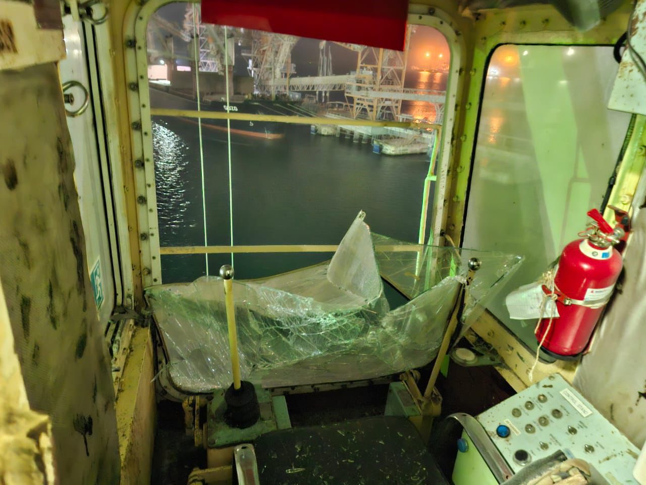 The interior of a control cabin of a vessel features a large front window that has been shattered and caved inward. Through the broken glass, a cargo ship and harbor infrastructure are visible at night. A red fire extinguisher and control panels remain in the debris-strewn workspace.