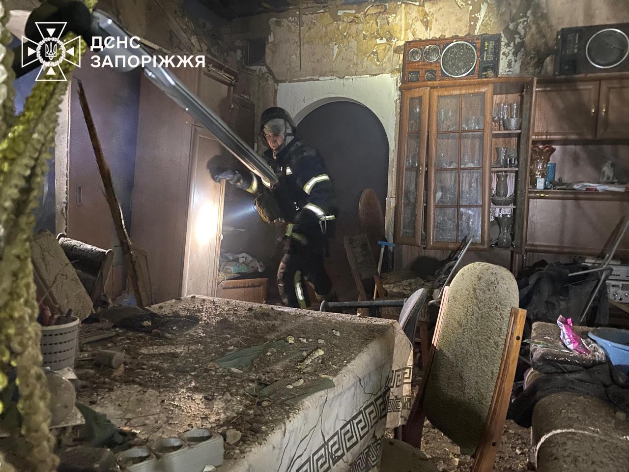 A Ukrainian rescuer from the State Emergency Service (DSNS) in Zaporizhzhia stands in the middle of a heavily damaged apartment. He is wearing a dark protective uniform with reflective stripes and a helmet, holding a bright flashlight to inspect the ruins. The room is devastated: wallpaper is peeling off the walls, a wooden cabinet with glass doors stands cracked, and every surface is covered in a thick layer of grey dust and debris. In the top-left corner, the official white logo of the DSNS Zaporizhzhia is visible.