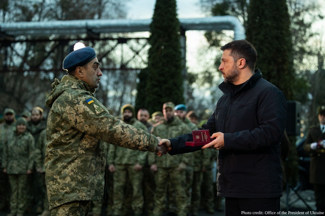 President Volodymyr Zelenskyy shakes hands with a Ukrainian soldier wearing a blue beret (identifying him as a member of the Air Assault Forces) and pixelated camouflage uniform while presenting him with a state award in a small red case. The ceremony took place outdoors in Kyiv on March 2, 2026, as part of the annual state Iftar event honoring Muslim military personnel and other defenders of Ukraine.