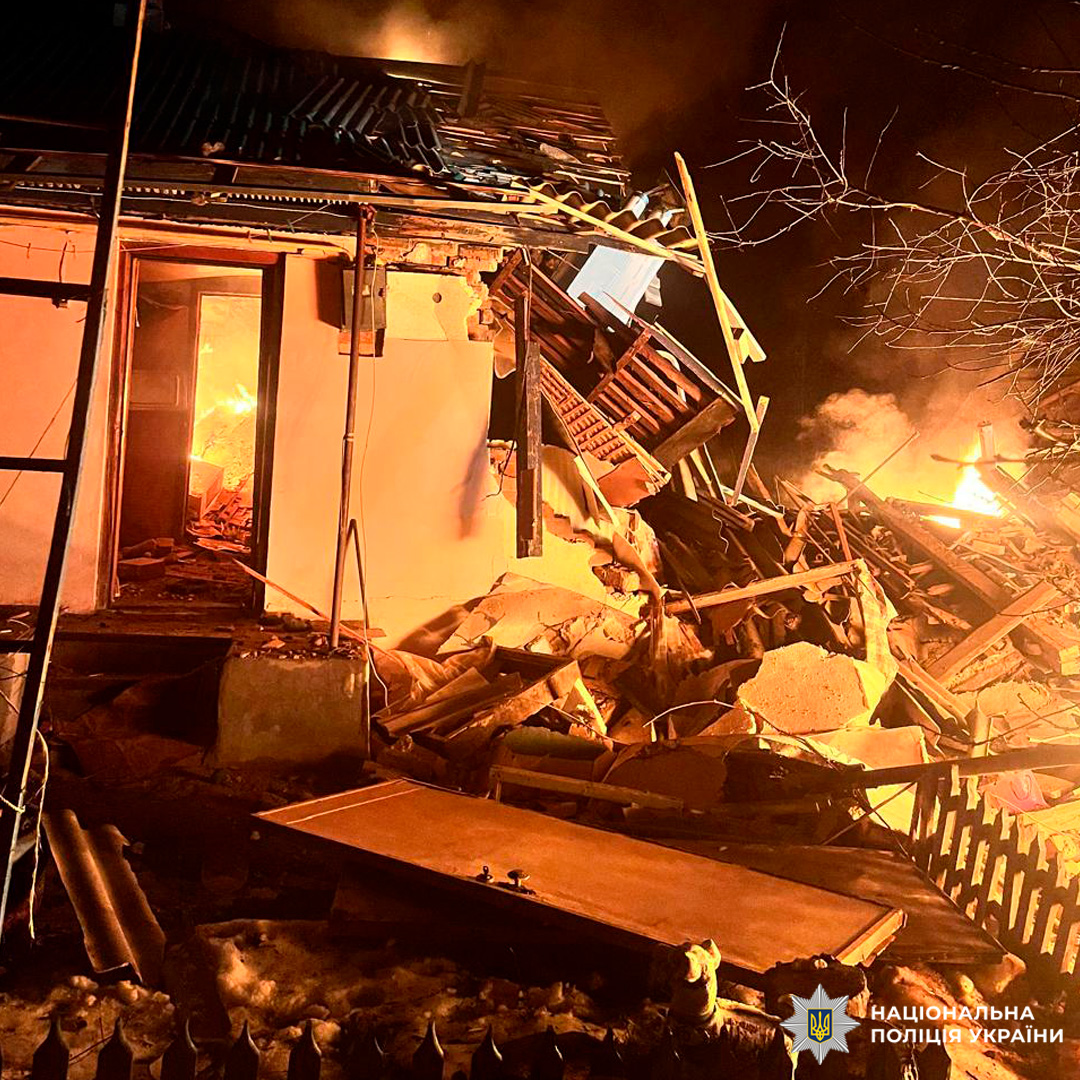 A small house is partially collapsed and engulfed in flames at night, with a doorway revealing a fire burning inside and debris scattered across the yard. The National Police of Ukraine logo is visible in the bottom right corner.