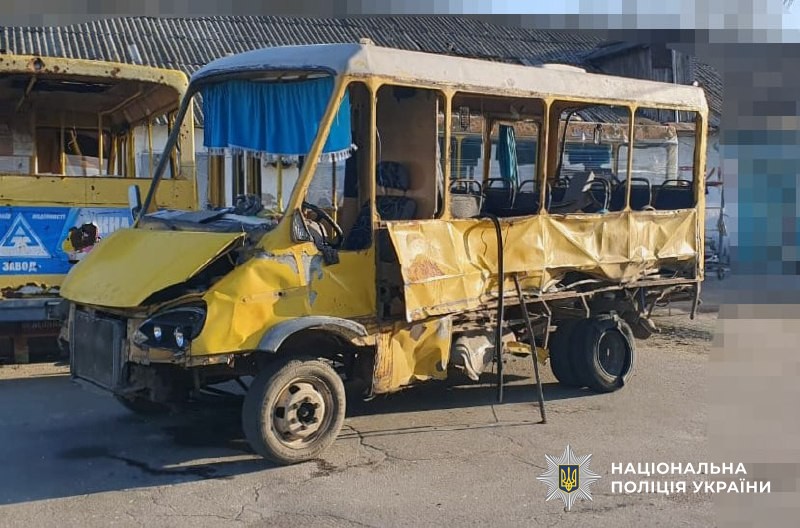 A yellow bus stands severely damaged with shattered windows, a damaged front end, and a shredded rear tire on an asphalt surface. The National Police of Ukraine logo is visible in the bottom right corner.