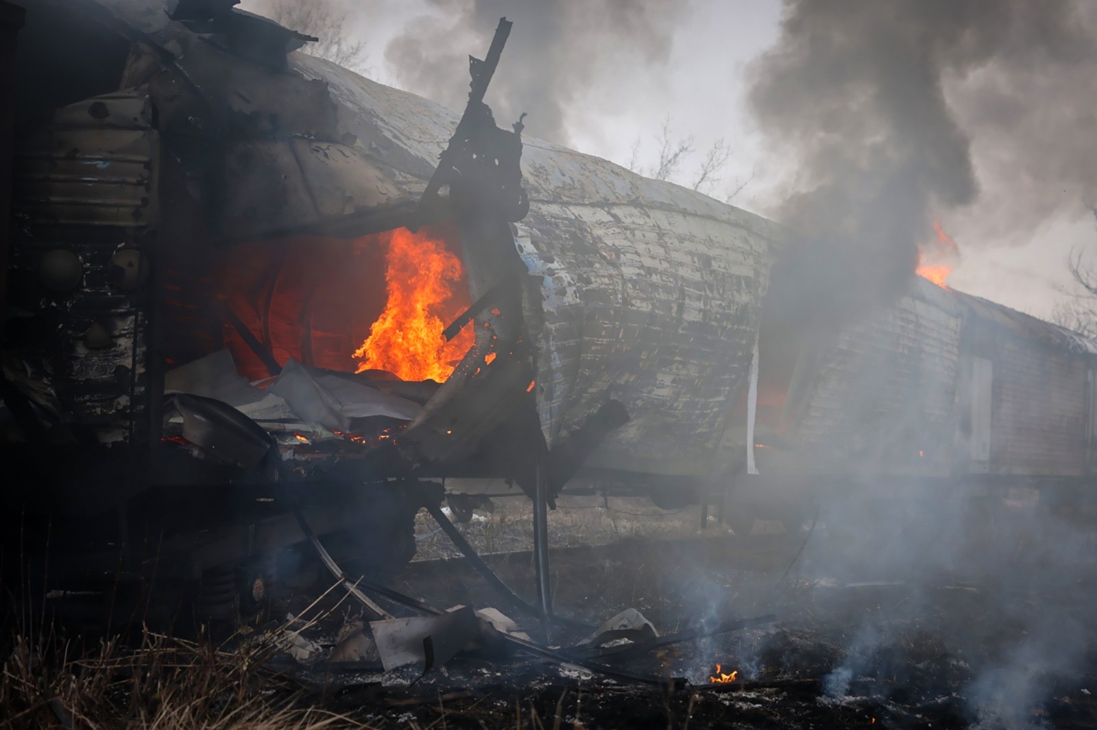 A severely damaged train car is engulfed in bright orange flames, with thick grey smoke billowing from a large hole in its side. The surrounding ground is scorched and littered with debris from the wreckage.
