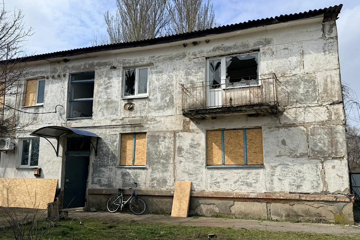 A two-story, peeling white-painted concrete residential building is heavily damaged. All ground-floor windows are boarded up with plywood. A central first-floor balcony has a broken door and window, with large shards of glass visible in the frame. Plywood boards rest against the wall near the entrance. A bicycle is parked outside. The sky is bright but partially cloudy, with tall trees in the background.