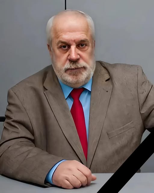 A formal memorial portrait of a senior man with grey hair and a neatly trimmed grey beard. He is wearing a light brown suit jacket, a blue shirt, and a solid red tie. He has a calm, thoughtful expression and is seated behind a grey desk. A black mourning ribbon is diagonally placed across the bottom right corner of the image.