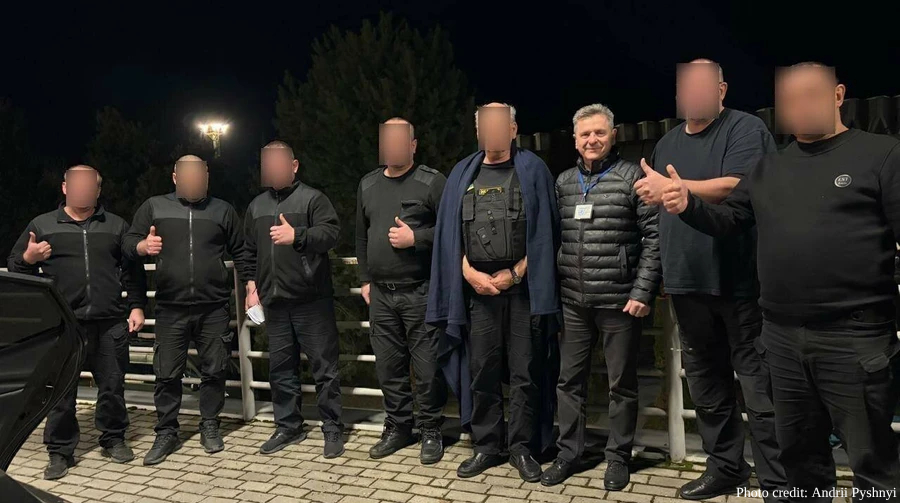 A group of nine men stands in a line outdoors at night on a paved surface. Most of the men have their faces blurred; several on both ends of the line are giving a "thumbs up" gesture.

In the center, a blurred man wears a tactical vest with a dark blue blanket draped over his shoulders. Second from the right, a man with grey hair, wearing a black puffer jacket and a lanyard, is the only one whose face is clearly visible. The background shows trees and a bright light in the distance. A watermark in the bottom right corner reads "Photo credit: Andrii Pyshnyi."