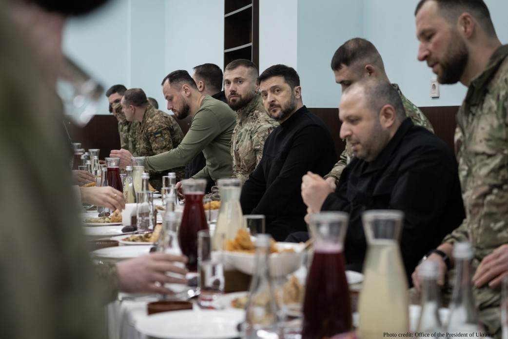 President Volodymyr Zelenskyy, Ukraine's security council head Rustem Umerov, and several Ukrainian soldiers in camouflage uniforms are seated at a long table during an Iftar ceremony in Kyiv on March 2, 2026. Zelenskyy looks directly at the camera while Umerov and the soldiers engage in conversation. The table is set with pitchers of juice and bowls of traditional food.