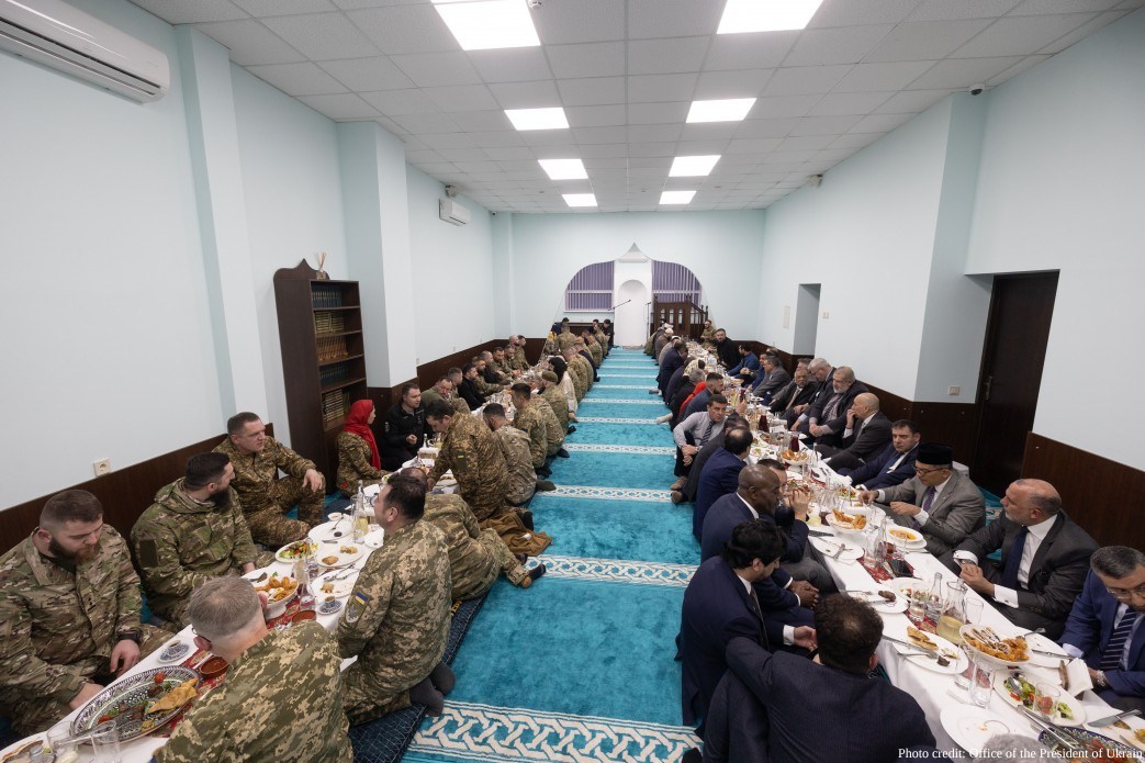 A wide-angle interior view of the Iftar ceremony in Kyiv on March 2, 2026, shows two parallel rows of participants seated at long tables spread across a turquoise-carpeted prayer hall. President Zelenskyy and high-ranking officials are seated on the right, while military personnel and religious leaders occupy the left, participating in the traditional meal to break the Ramadan fast.