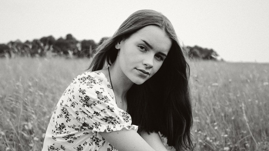 A black-and-white portrait captures a young girl with long hair wearing a floral-patterned dress while sitting in a field.