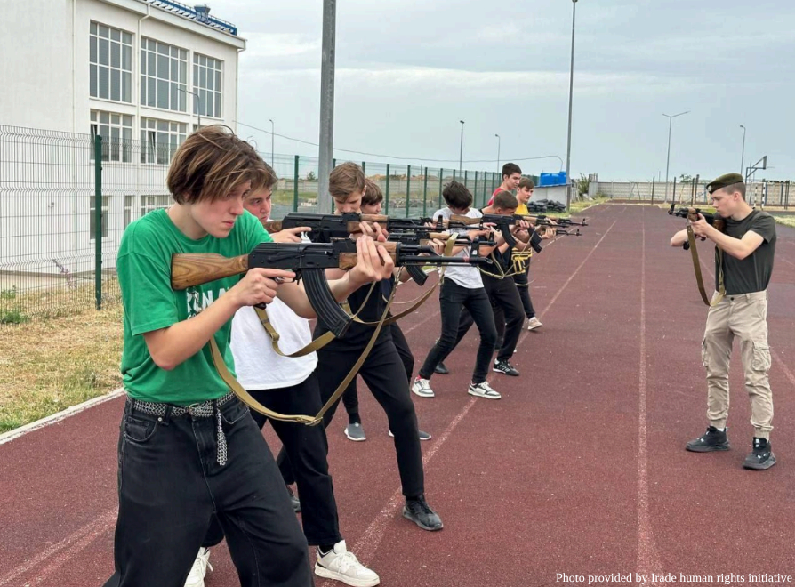 A line of seven teenagers stands on a red athletic running track, each holding a Kalashnikov-style assault rifle in a shooting stance, aiming toward the right. On the far right, a young man in a dark t-shirt and a military-style green beret holds a similar rifle, appearing to lead or demonstrate the drill. The group is positioned in front of a white multi-story building and a tall wire fence. A watermark in the bottom right corner reads: "Photo provided by Irade human rights initiative."