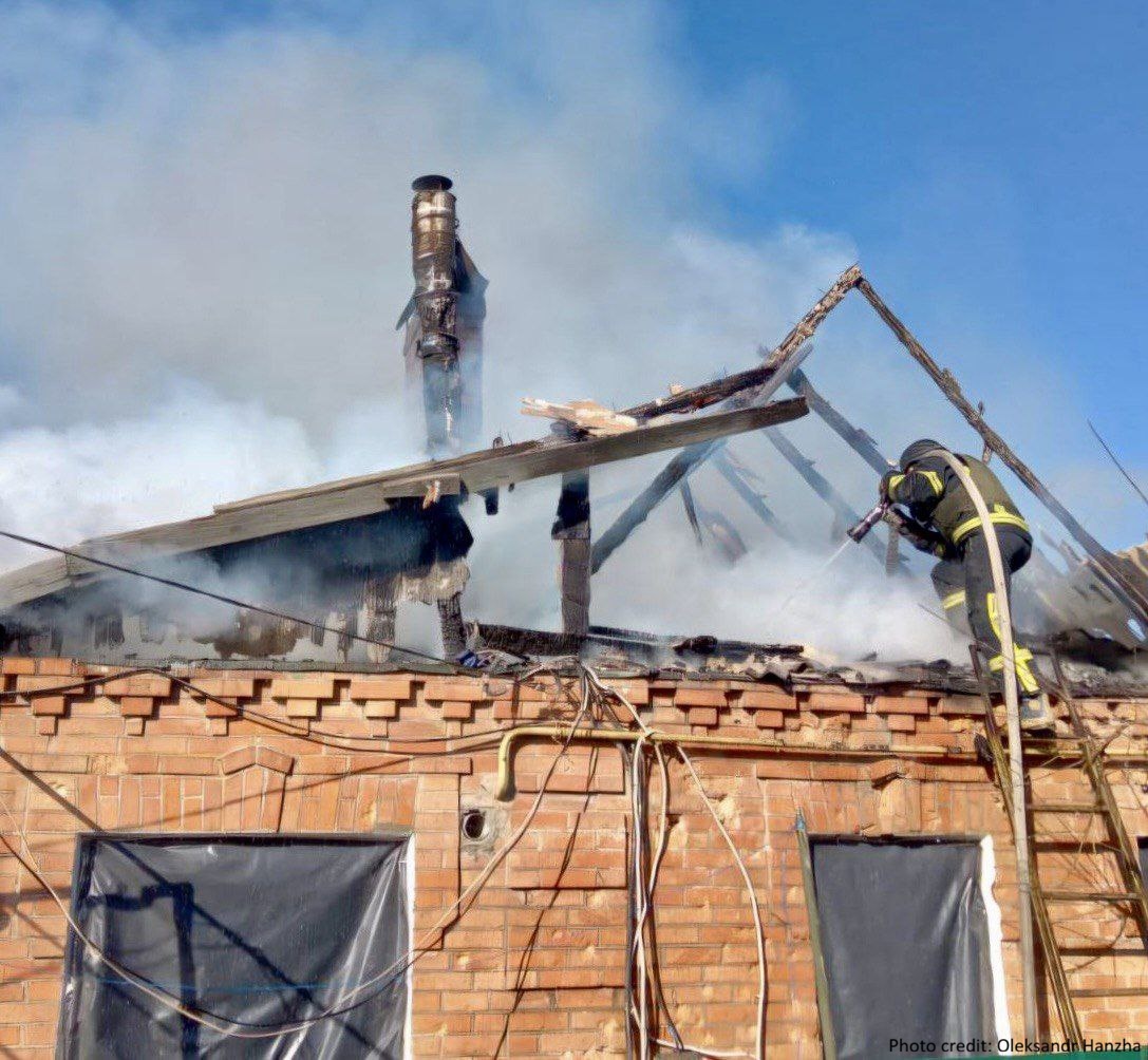 A firefighter stands on a ladder and directs a water hose toward the charred, smoking skeleton of a roof on a brick building. The windows below are covered with dark plastic, and a photo credit for Oleksandr Hanzha is visible in the bottom right corner.