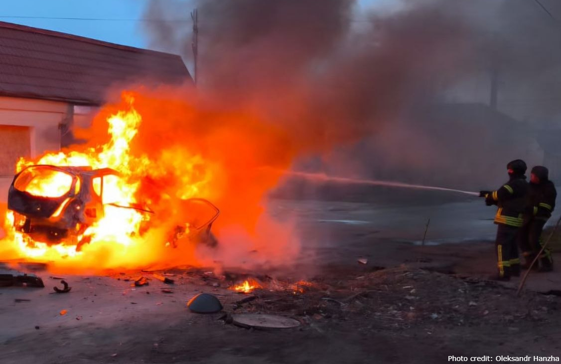 A car is completely engulfed in intense orange flames and thick black smoke on a street during twilight. To the right, two firefighters in protective gear direct a high-pressure stream of water toward the blaze. Debris is scattered across the pavement in the foreground. A watermark in the bottom right corner reads "Photo credit: Oleksandr Hanzha."