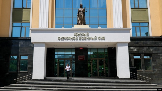 A statue of Lady Justice stands above the entrance of the Southern District Military Court in Russia, a modern building with large glass windows and dark stone pillars. A person descends the wide concrete steps leading to the main doors.