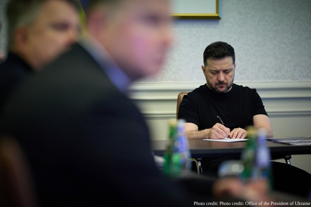 President Volodymyr Zelenskyy of Ukraine is seated at a long conference table, leaning forward and focused as he writes on a sheet of paper with a pen. He is wearing a black short-sleeved t-shirt. The foreground shows the blurred out-of-focus profiles of other officials attending the meeting. The background is a simple white wall with classic molding. A watermark at the bottom right attributes the photo to the Office of the President of Ukraine.