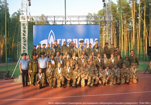 A large group of teenagers in military-style camouflage uniforms poses for a photo on an outdoor athletic track. Behind them is a massive blue billboard featuring the white Gazprom logo. Several adults in official uniforms stand among the youth. The setting is a wooded area with tall pine trees.